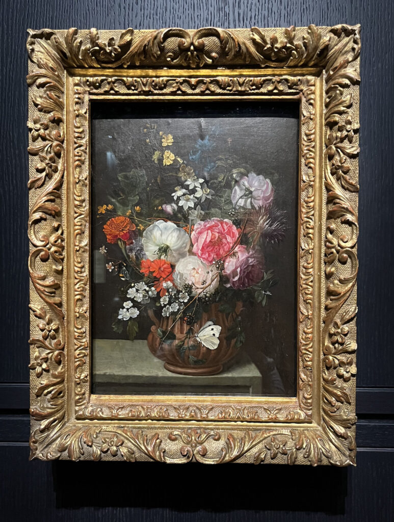 Still life floral oil painting attributed to Maria van Oosterwyck from the women artists show, featuring a variety of mostly pink, white and red flowers with a branch of black berries in a vase, with a white butterfly fluttering around the vase, all surrounded by an ornate gold frame.