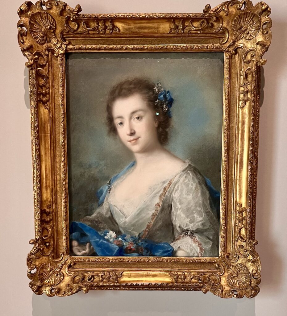 A pastel portrait in a gold frame by Rosalba Carriera of an 18th-century woman, gazing to her left at the viewer, and wearing a white lacy dress under a velvety blue cloak in which she cradles a half-dozen red, white and blue flowers.