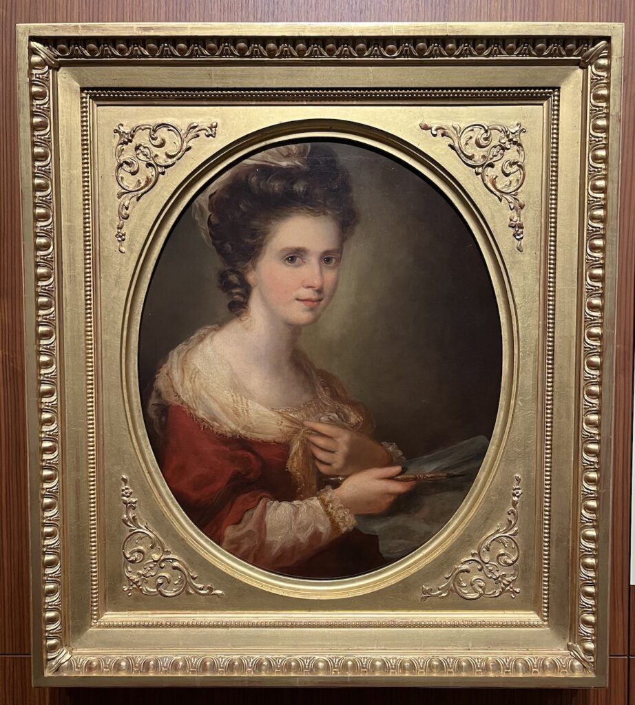 An oval portrait in a square gold frame of a young woman with thick brown hair, wearing a rust-colored dress with a lacy collar, holding a paintbrush in her right hand and holding the front of her dress with her left hand.