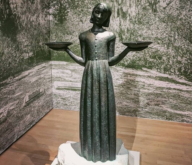 Photo of Judson's Bird Girl statue. The girl wears a long dress and holds a shallow dish in each hand, held out to her sides with her elbows bent such that her upper arms are parallel to her torso.