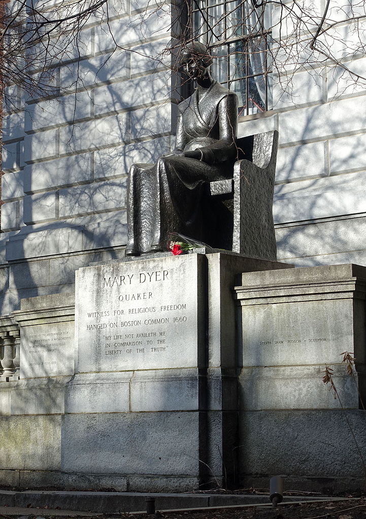 Photo of the Boston version of Sylvia Shaw Judson's statue of Mary Dyer, overlaid with tree branch shadows. A single red rose sits at the martyr's left foot.