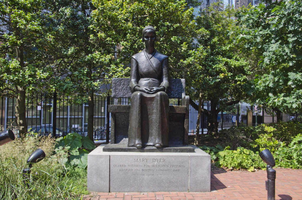 Photo of the Judson statue of Mary Dyer that sits on a red brick patio surrounded by trees and green ground cover.