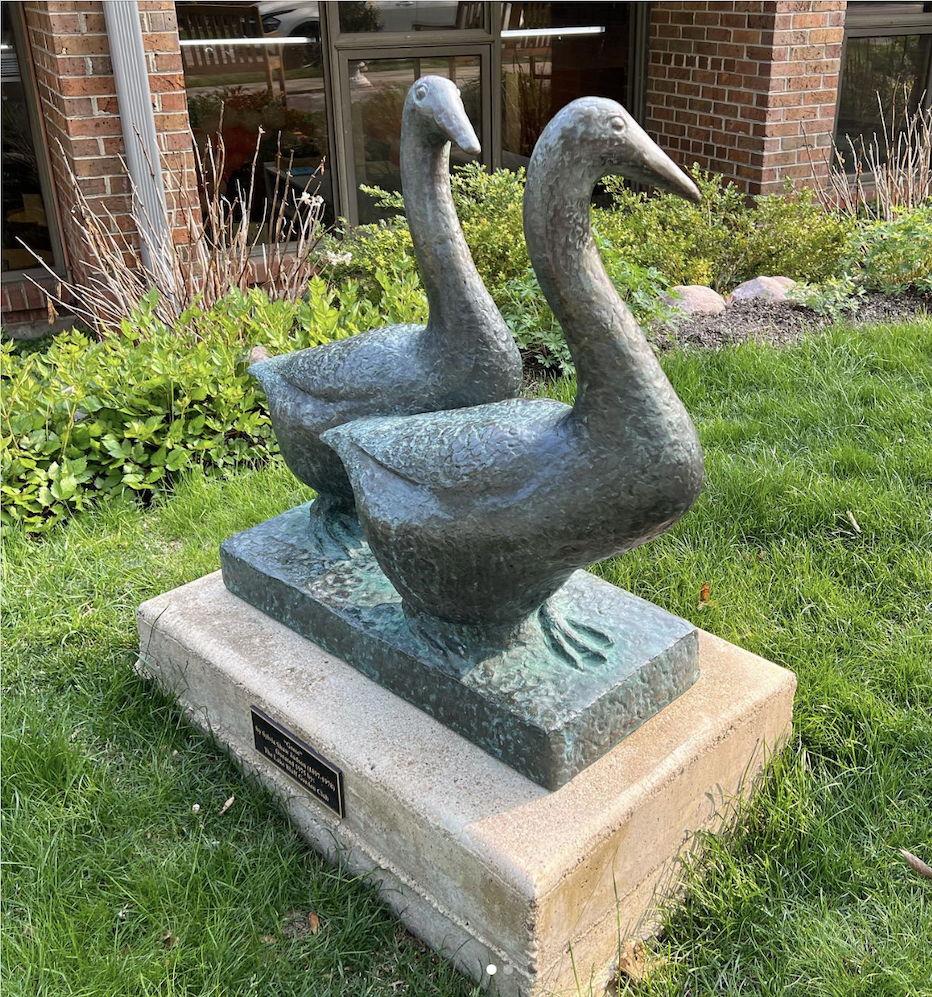 Statue of two Canada geese; sculpture is made from a metal and has developed a greenish patina.