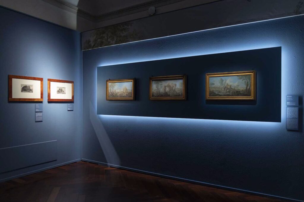 An installation view of two intersecting walls. On the wall at the viewer's left are two framed artworks, the content of which is not readily distinguished. On the perpendicular wall are three framed landscapes. 