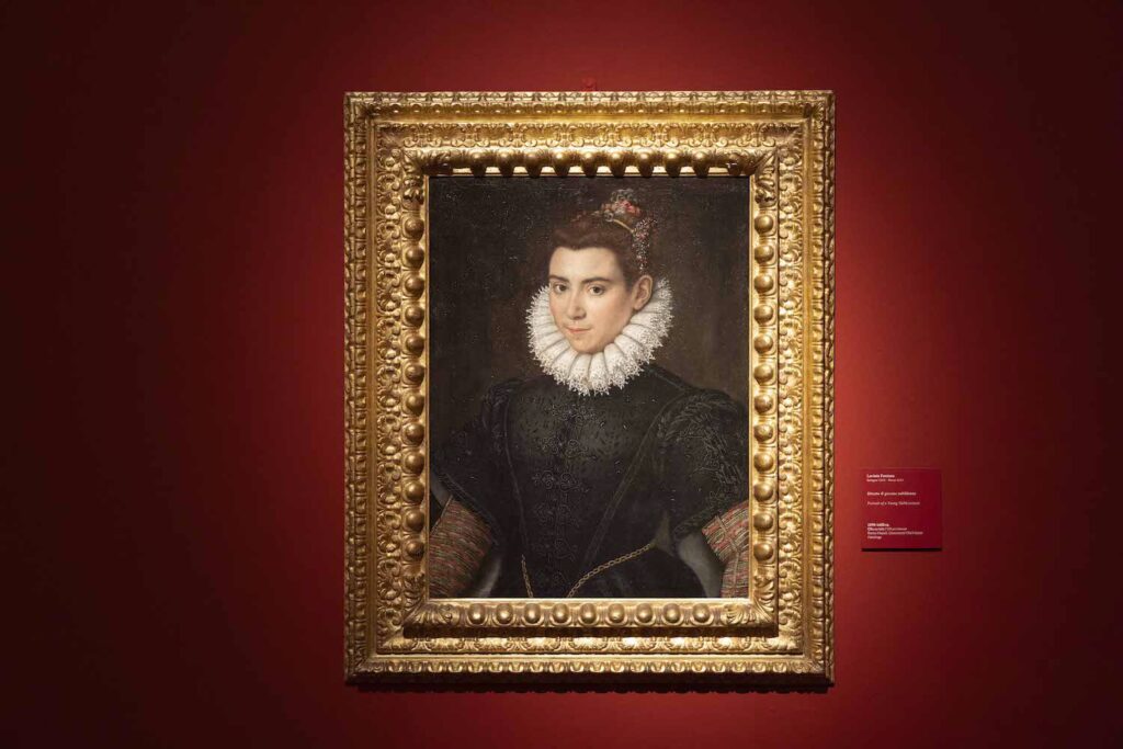 Installation view of a portrait of a woman in a black dress and a high white neck ruff. The painting is within an elaborate gold frame, and is hung on a dark red wall.