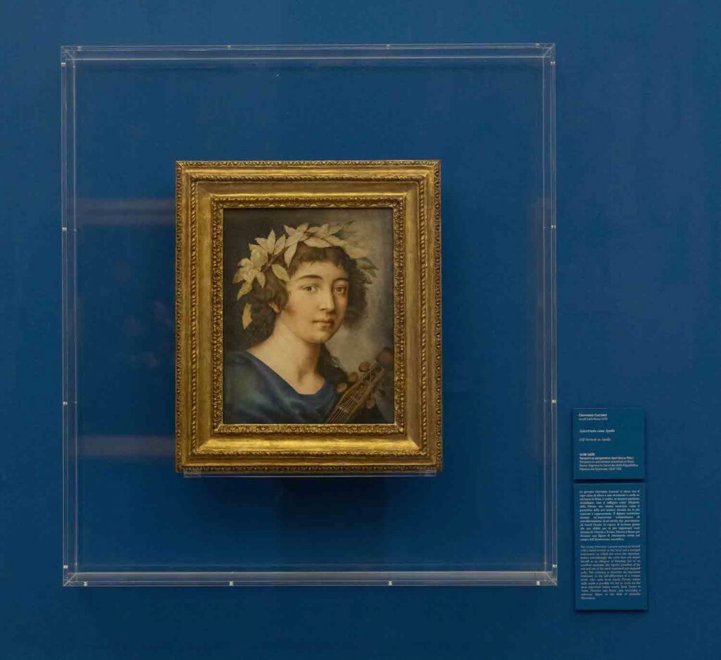 An installation view of a single paining behind glass or clear plastic. The smallish painting depicts the bust of a young woman with laurels in her hair. The neck of a musical instrument, perhaps a lute. angles across the lower right of the painting.