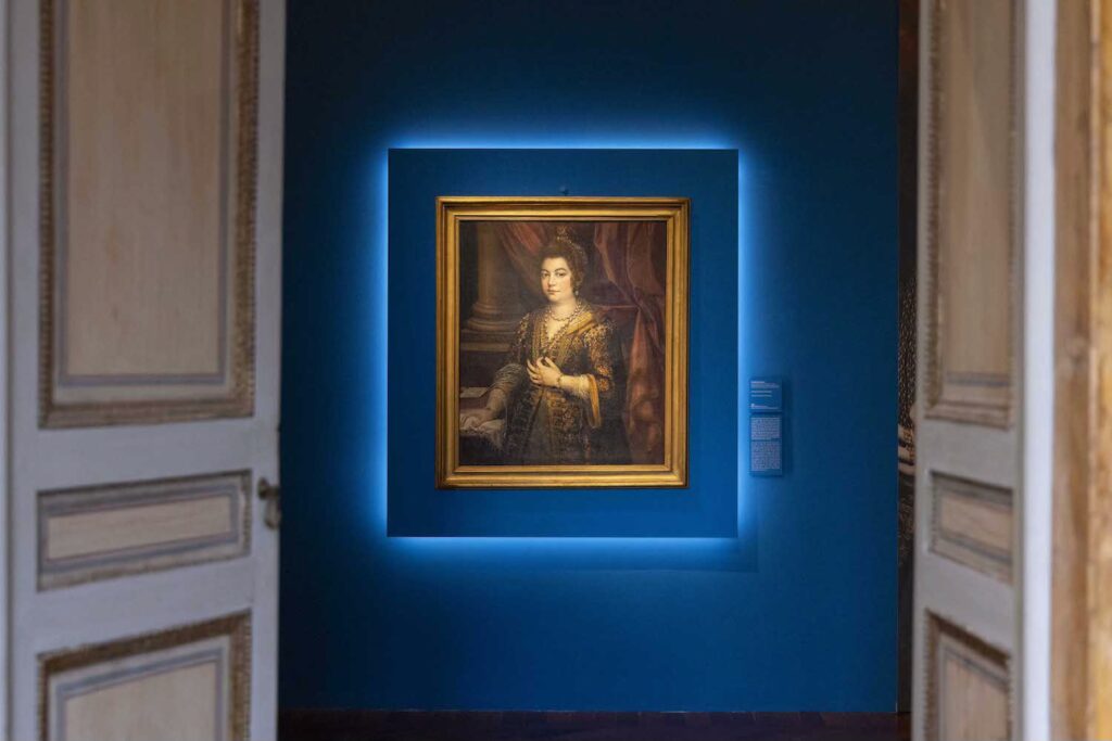 An installation view of a single painting, a portrait of a woman. The framed artwork is hung on a wall; there is lighting around the frame. At either side of the photo are two white and gold doors, opening into the room that contains the painting.